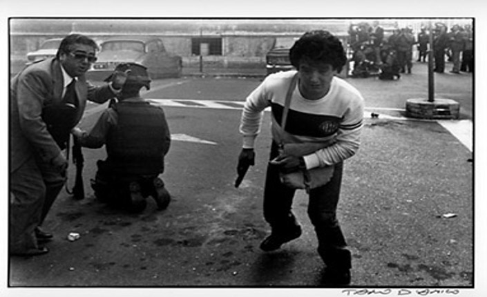 Foto di Tano D'amico raffigurante l'agente Giovanni Santone in borghese armato durante gli scontri. Alla sua destra un funzionario e un agente in divisa